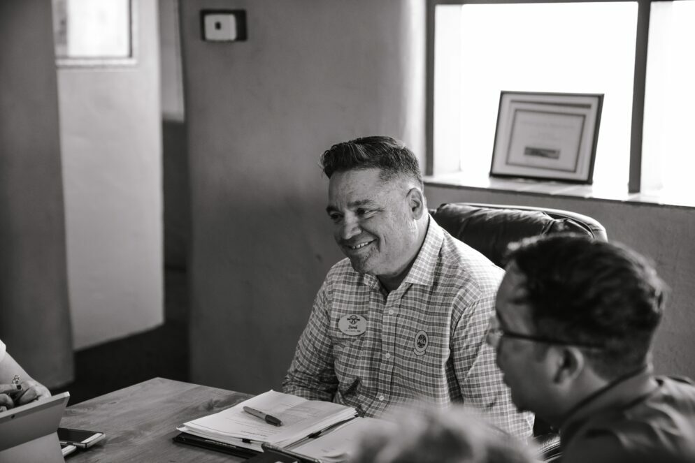 A white man with short dark hair sits at the head of a table during a meeting. He is smiling, with his hands on the arm of the chair, looking at someone across the table (not pictured). He has a portfolio open in front of him, with a pen on top. Two people sit on the side of the table to his left. The photo is black and white.