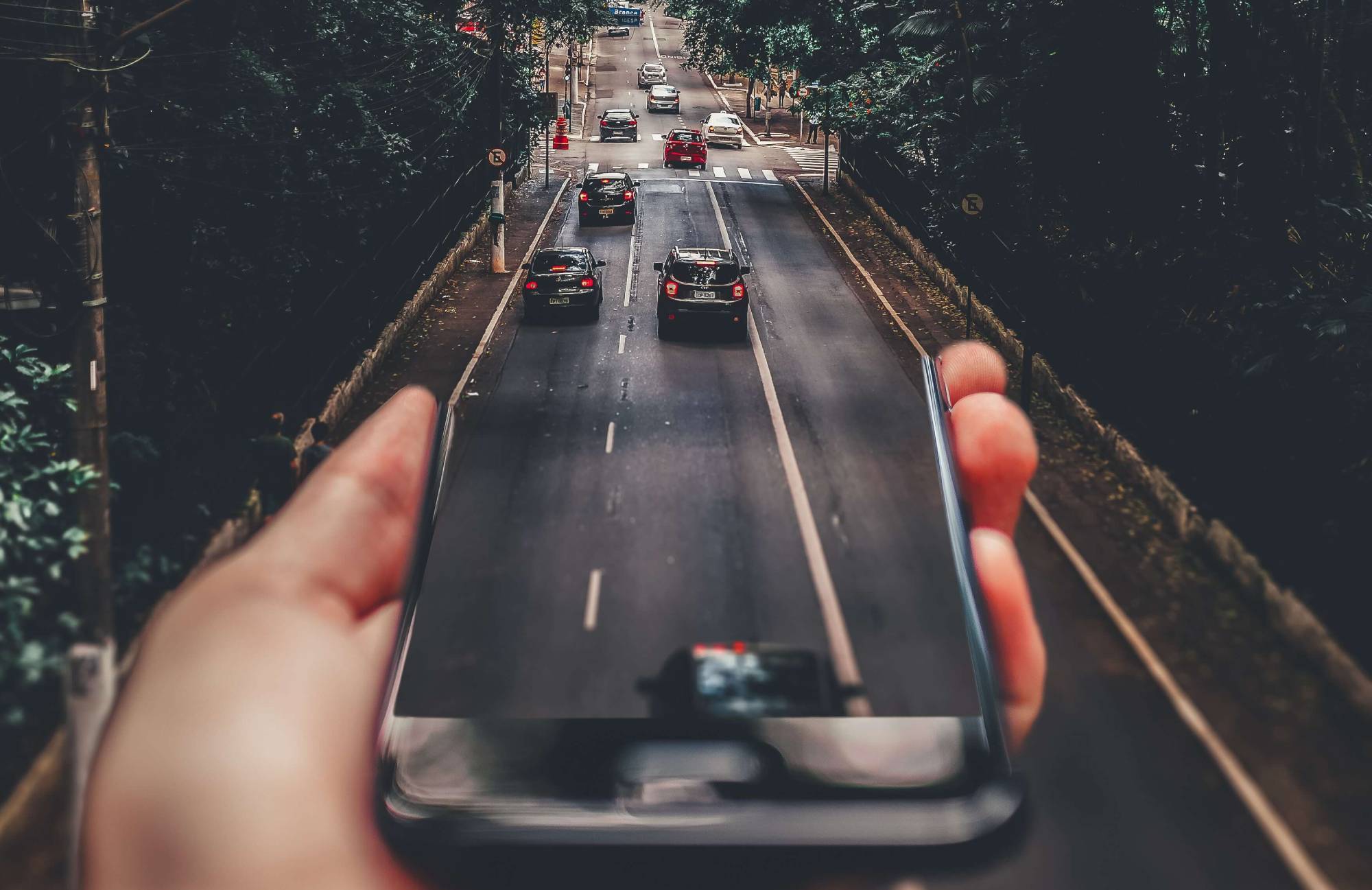 Several cars drive along a street, which blends into the screen of a mobile phone being held up by a white left hand. It gives the impression that the content of the phone and the real world have blended.
